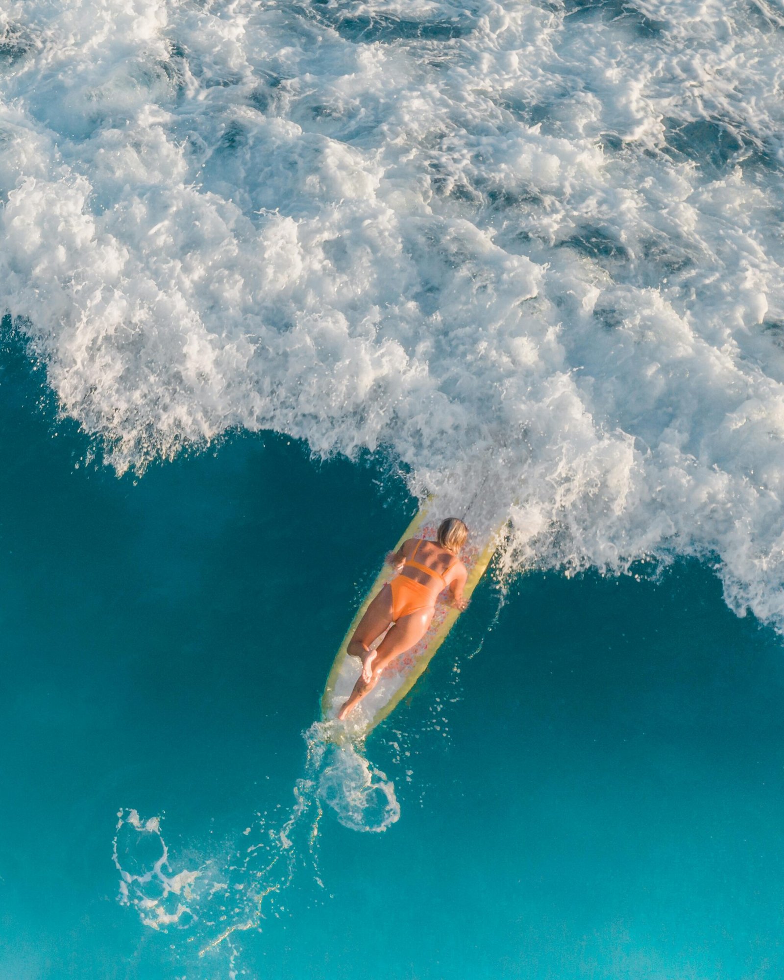 Aerial view of a woman surfing a wave in the ocean, showcasing clear blue water and foamy waves.